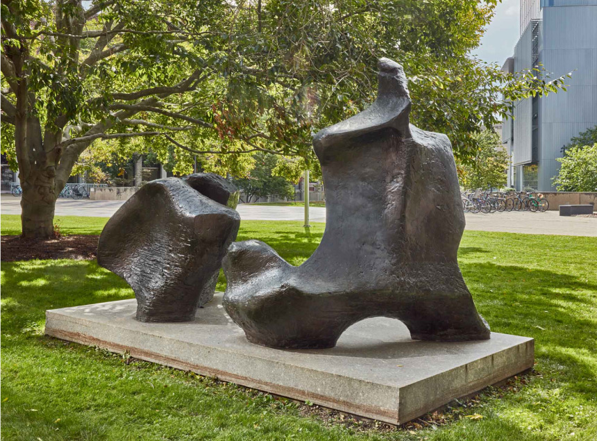 An abstract bronze sculpture of various natural shapes sits on top of a pedestal in a grassy lawn.