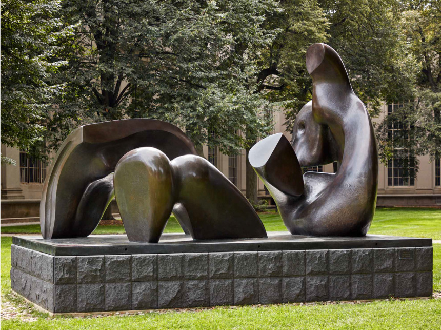 An abstract bronze sculpture of various natural shapes sits on top of a pedestal in a grassy lawn.