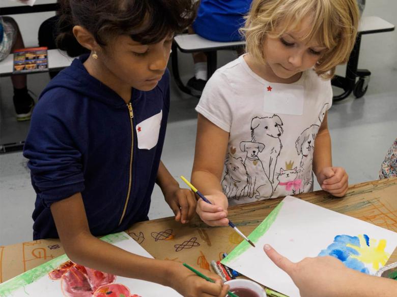 Two kids at a table, holding paintbrushes, painting with watercolors