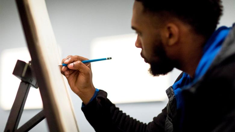 Man leaning in towards easel, drawing on paper with blue colored pencil