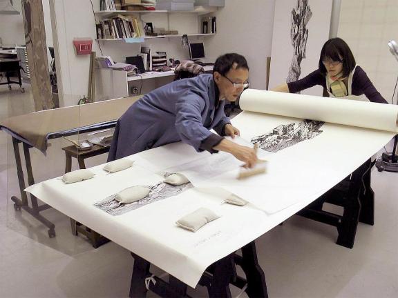 Conservator leaning over and brushing scroll, while another conservator holds on to rolled-up end of scroll