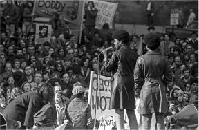 Two figures wearing trench coats stand on a stage and look out at a large crowd.