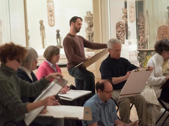 Man drawing on sketchpad amongst seated artists in gallery
