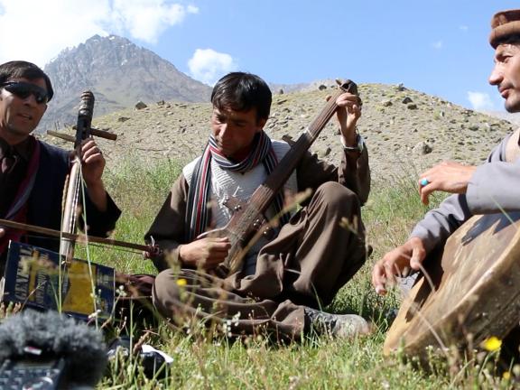 Three Wakhi musicians play their instruments in front of a mountain in a summer pasture