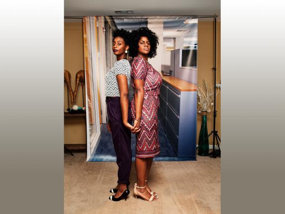 Color photograph of two young women standing back to back looking into the camera in front of a backdrop of an office 