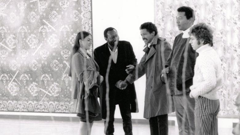 Four people stand in a line against a white wall with large tapestries against it. Black-and-white image.