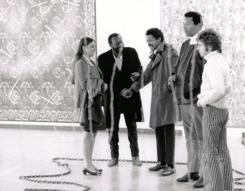 Four people stand in a line against a white wall with large tapestries against it. Black-and-white image.