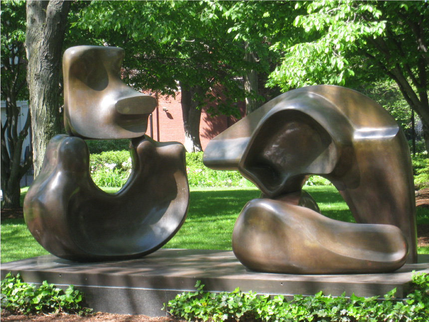 An abstract bronze sculpture of various natural shapes sits on top of a pedestal in a grassy lawn.