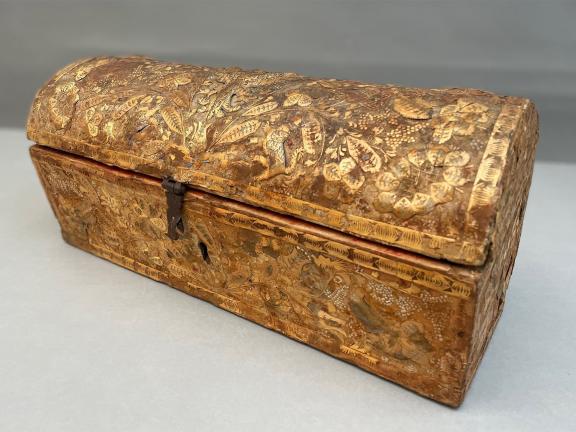 A photo of a long, rectangular, casket decorated with golden straw designs and covered with a domed, hinged top.
