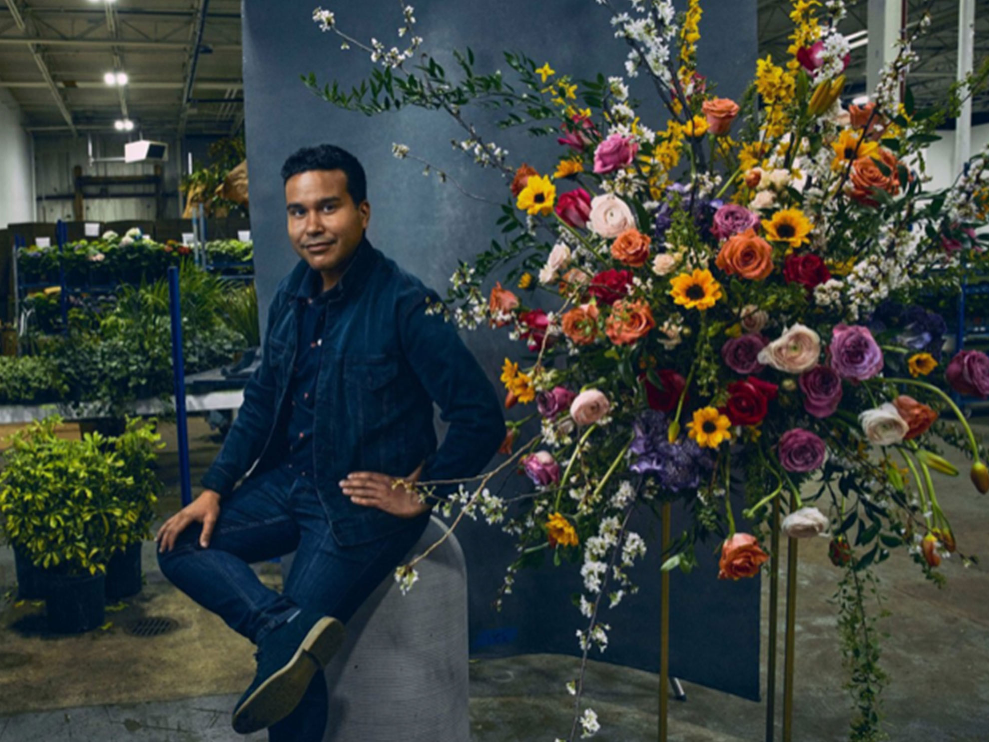 A man sits on a stool next to a large colorful floral arrangement
