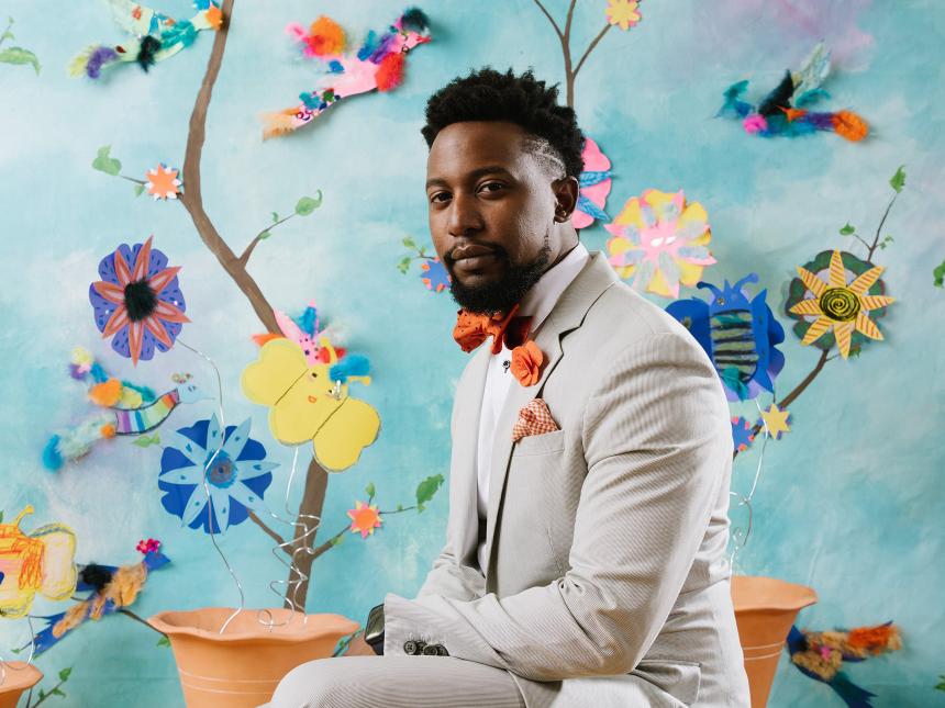 portrait of a man in suit and red bow tie sitting in front of backdrop featuring flowers, leaves and branches made of construction paper