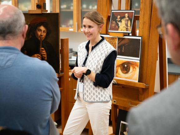 Conservator talking in front of paintings and printed details of paintings, displayed on easels; talk attendees in foreground