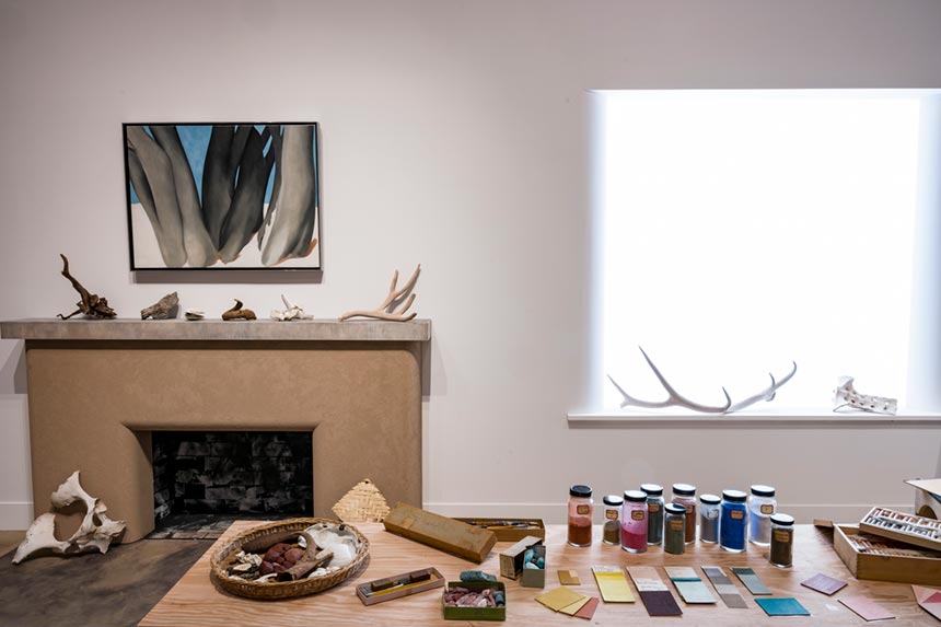 A spare studio space with animal bones on the mantle, a simple painting above a fireplace, and a cluttered table of paint vials.