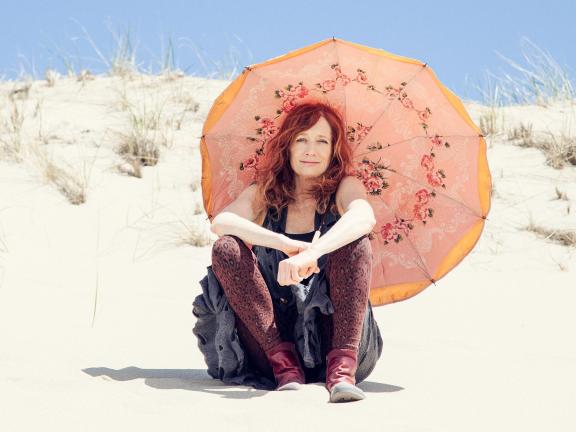 Musician Patty Larkin holds a colorful parasol and sits on the beach. 