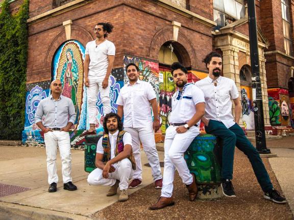 Press photo of the band Quitapenas: a band of six men posing in front of a brick wall with colorful murals. 