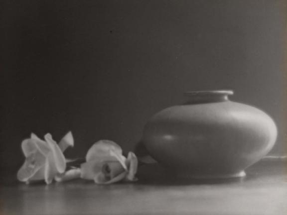 A still life black-and-white photograph of a squat vase beside two white roses resting on a table.