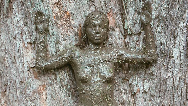 A woman covered in mud stands with her arms up and back against a tree.