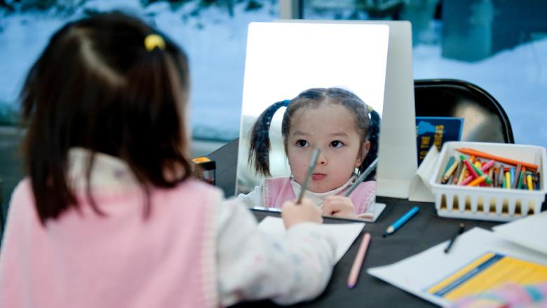 Child with pigtails sitting at table in front of mirror, holding colored pencil to draw on paper