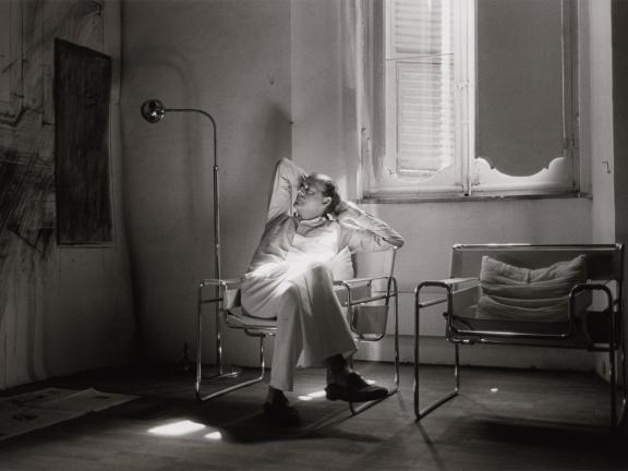 A photograph of artist Cy Twombly sitting in a chair by an open window with his legs crossed and his arms supporting his head 