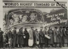 People stand in a line, some holding baskets, bags, or pails, in front of a billboard.