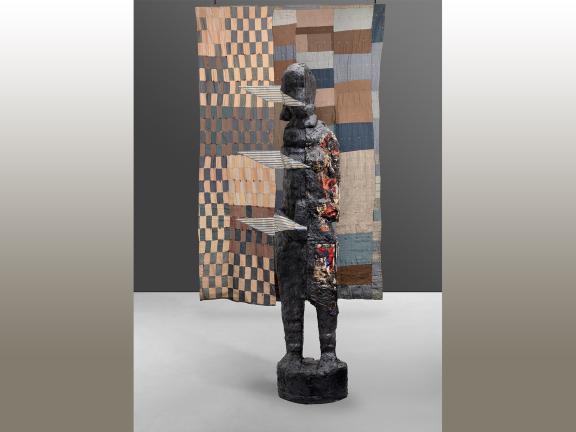 Photograph of a large sculpture of a human form in front of a quilt.