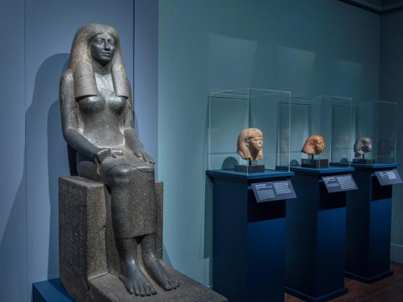 ancient Egyptian statue of a woman sitting against a blue wall next to cases with sculptures of heads.
