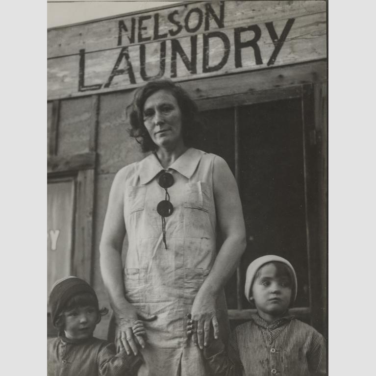A woman, Mrs. Nelson, stands in front of her laundry business holding the hands of her two young children.