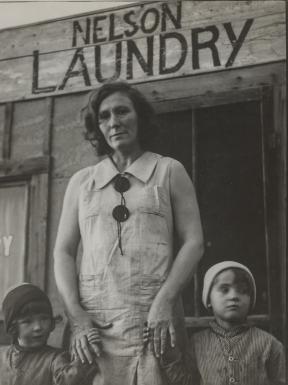 A woman, Mrs. Nelson, stands in front of her laundry business holding the hands of her two young children.