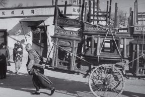 A man pulls along various pieces of wooden furniture on a wheeled cart. 