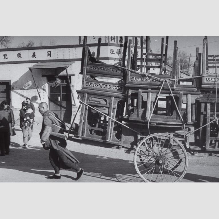 A man pulls along various pieces of wooden furniture on a wheeled cart.