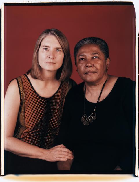 Two individuals sit closely next to each other and pose for a photo against a red background.