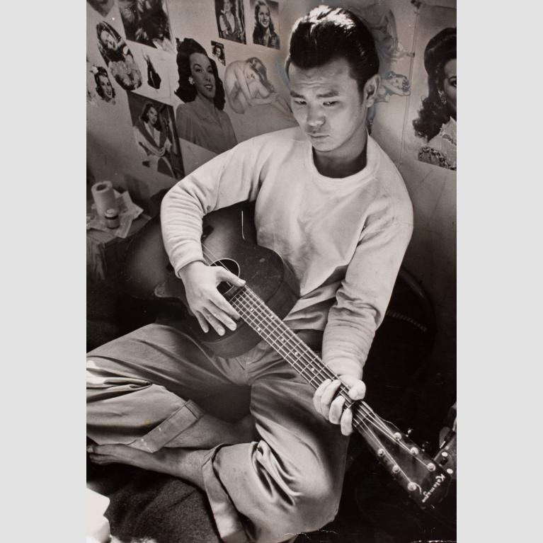 A young man, Carl Mydans, sits cross-legged and plays guitar.