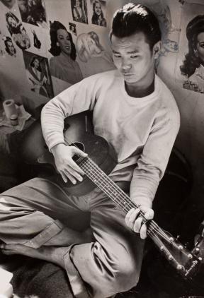 A young man, Carl Mydans, sits cross-legged and plays guitar. 