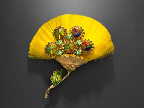 A flower brooch consisting of yellow silk fibers, a gilded metal stem, and green enameled leaves.