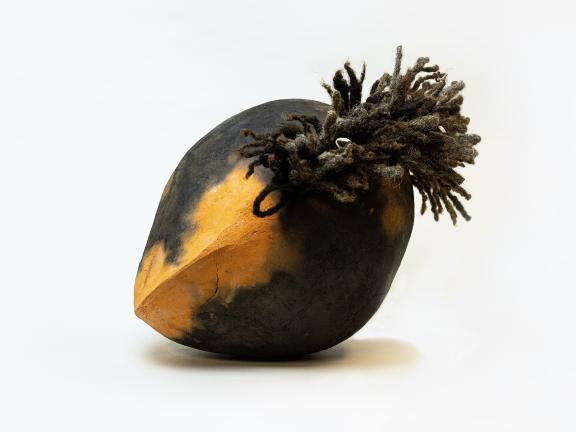 A photograph of a ceramic work made from plantation funerary soil, shaped into an oval sphere and implanted with locks of real human hair.