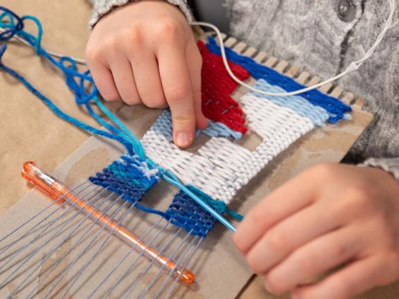 A person whose hands only are visible weaves a blue and white pattern on a small loom resting on a table.