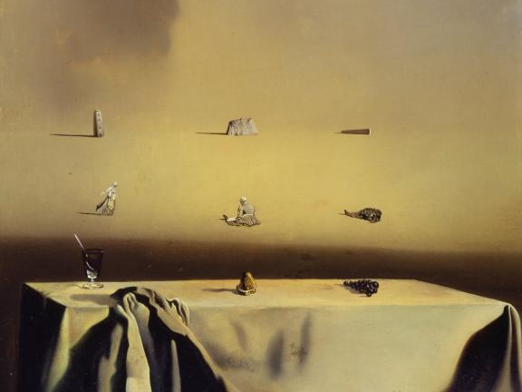 In the foreground, a group of grapes, a spoon in a glass, and a gold object sit atop a table covered in a scrunched white tablecloth; the background is dotted with small women and stone structures. 