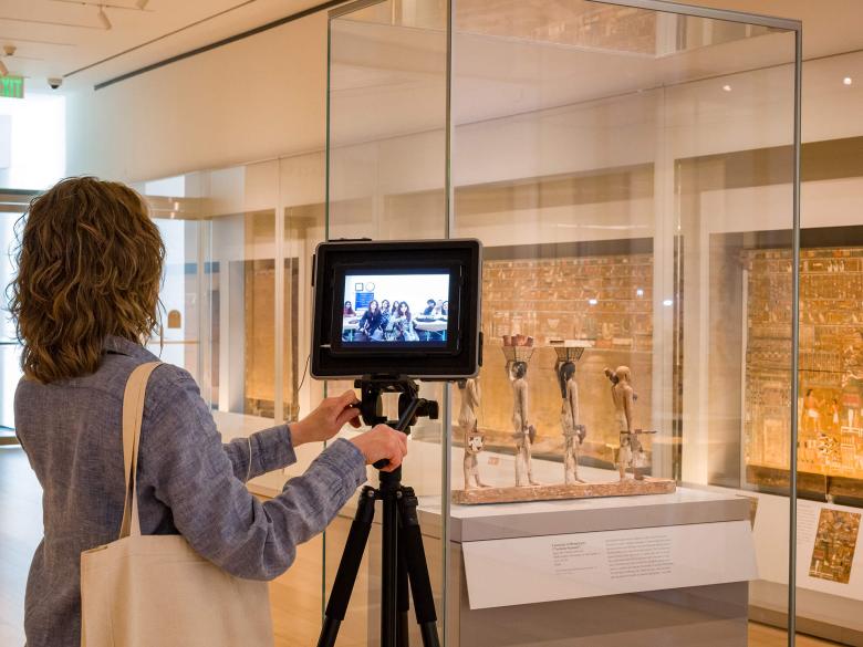 An MFA instructor operating a camera on a tripod pointed wooden Egyptian sculptures; panels of hieroglyphs in background
