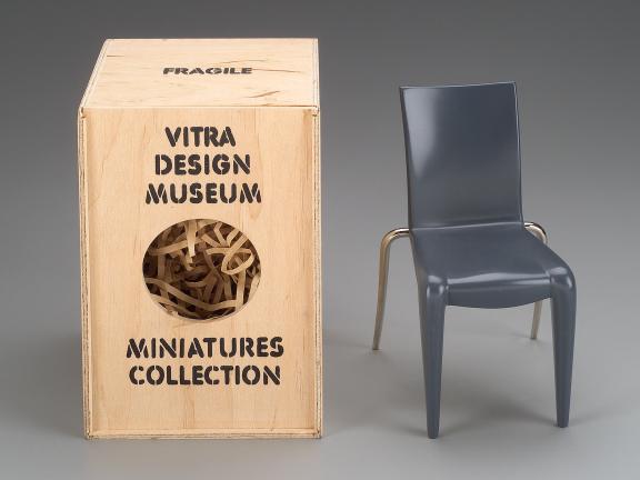 A miniature chair stands next to it's miniature shipping container that reads: "Fragile. Vitra Design Museum. Miniatures Collection."