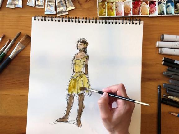 Overhead view of drawing pad on tabletop, with artist's hand painting in drawing of ballerina