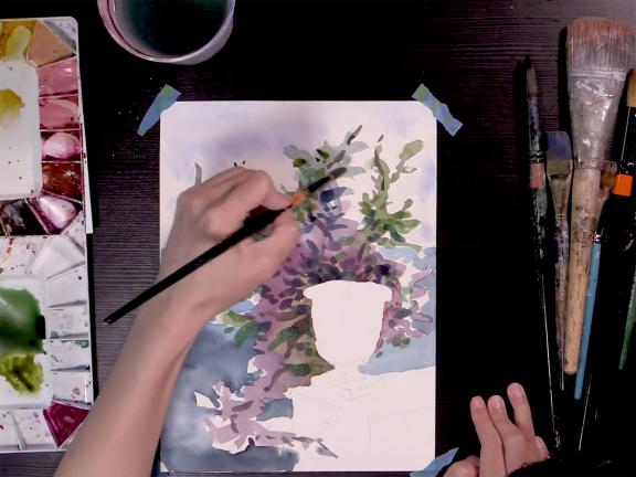 Overhead view of table, with hand holding paintbrush, painting with watercolors on sheet of paper