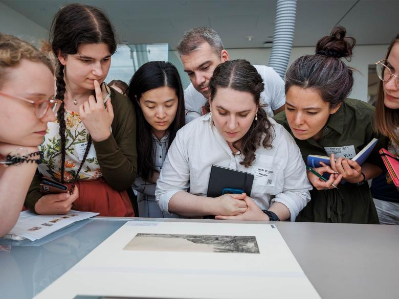 A group of people gather around a print displayed face up on a tabletop.