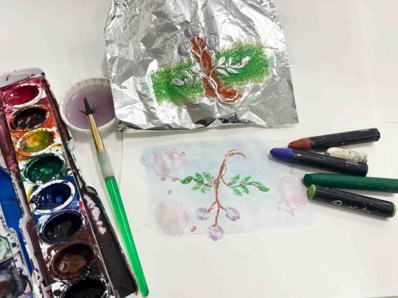 Various art supplies gathered on a table covered in white paper: watercolors, crayons, embossed foil, and an in-process dry print.